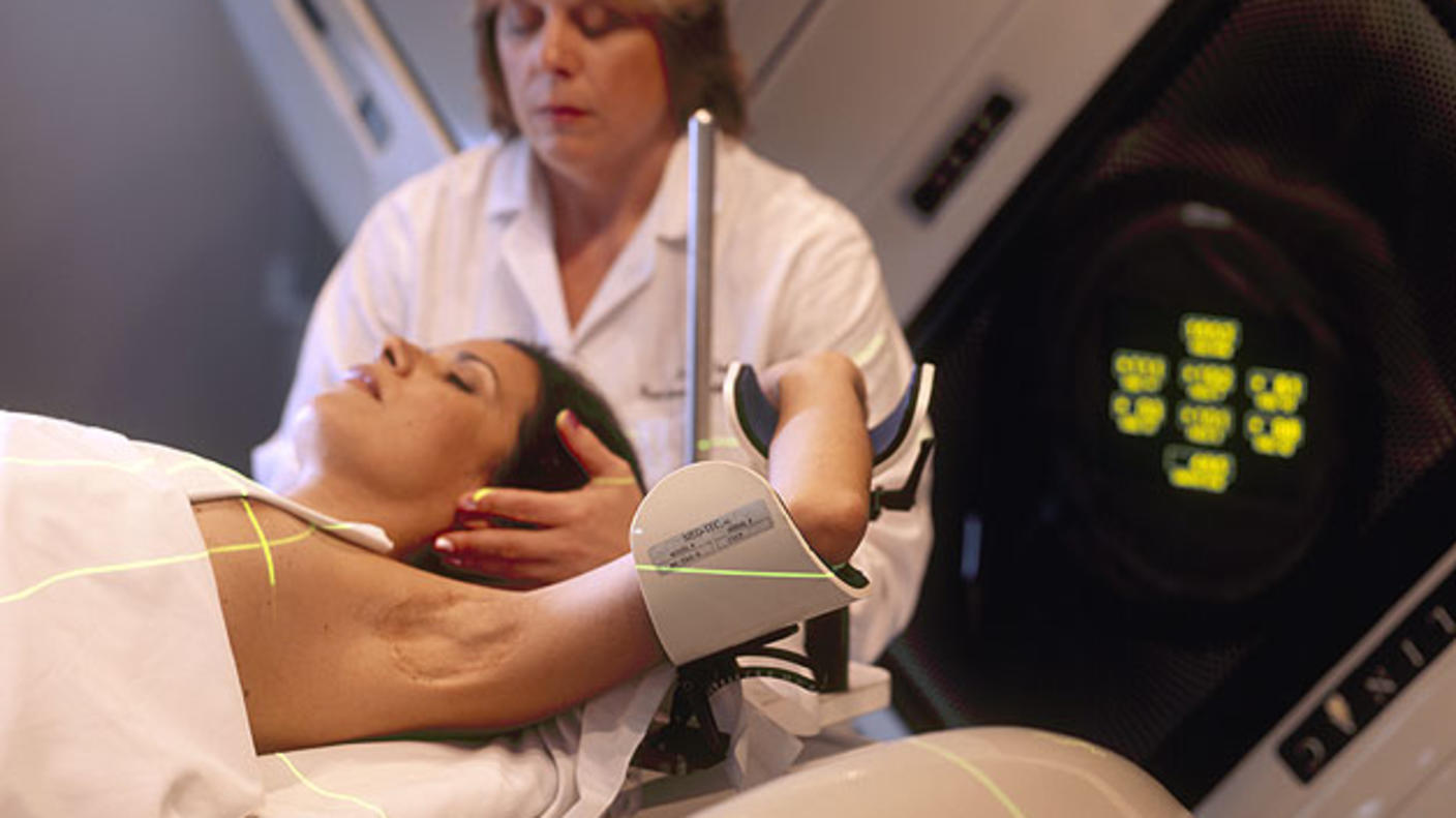 A Hispanic woman (lying on her back) is being prepared for radiation therapy by a Caucasian female radiation therapist.